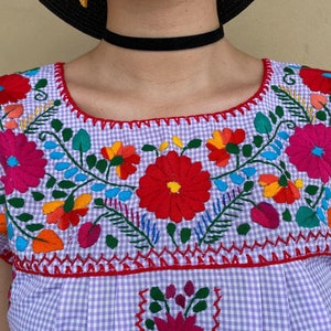 Peut inclure: Une blouse brod&eacute;e en gingham violet et blanc avec des motifs floraux color&eacute;s. La blouse a un col rond et des manches courtes.