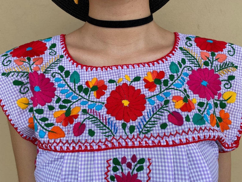 Peut inclure: Une blouse brod&eacute;e en gingham violet et blanc avec des motifs floraux color&eacute;s. La blouse a un col rond et des manches courtes.