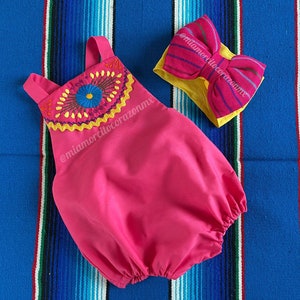 Op de afbeelding: Een roze baby-romper met een kleurrijk geborduurd bloemetjespatroon en een roze en gele strik. De romper ligt op een blauw-wit gestreept kleed.