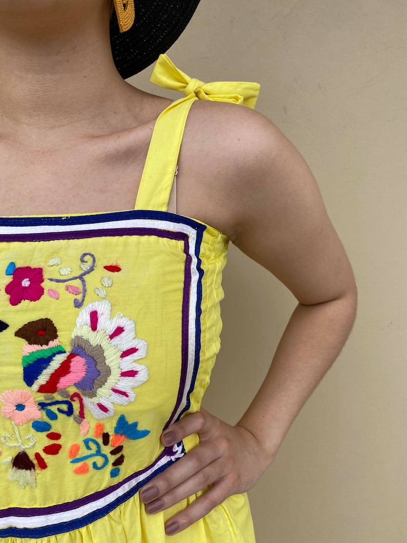 May include: Yellow sleeveless dress with colorful embroidered flowers and a bow on the strap.