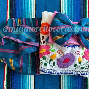 Op de afbeelding: Een collectie handgemaakte babyartikelen, waaronder een teal en roze gestreepte hoofdband, een bijpassende gerimpelde luierbroek en een wit geborduurd topje met bloemmotieven. De items worden gepresenteerd op een blauw gestreept textiel.