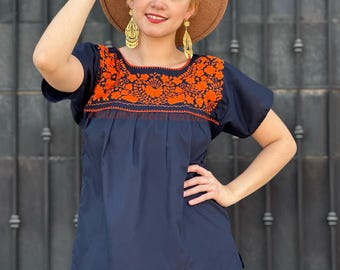 May include: A navy blue embroidered blouse with orange floral embroidery. The blouse has short sleeves and a round neckline. The model is wearing a brown hat and gold earrings.