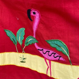 May include: A pink flamingo embroidered on a red fabric background. The flamingo is standing on a yellow strip with green leaves and a small plant.