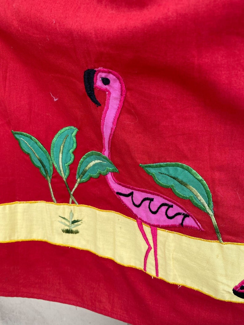 May include: A pink flamingo embroidered on a red fabric background. The flamingo is standing on a yellow strip with green leaves and a small plant.