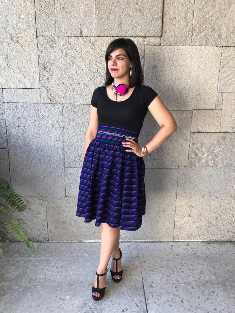 May include: A woman wearing a black short-sleeved shirt with a pink and blue necklace. She is wearing a black and purple striped skirt with a black waistband that says "@miamordilecerrania.mx". She is wearing black heeled sandals.