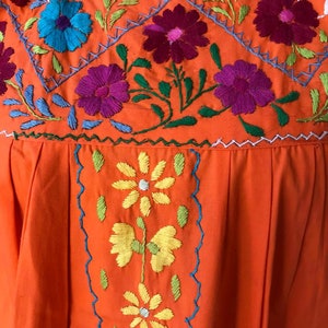 May include: Orange cotton dress with colorful floral embroidery. The embroidery features a variety of flowers in shades of pink, blue, yellow, and green.
