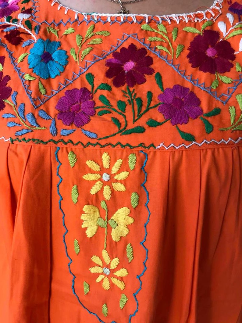 May include: Orange cotton dress with colorful floral embroidery. The embroidery features a variety of flowers in shades of pink, blue, yellow, and green.