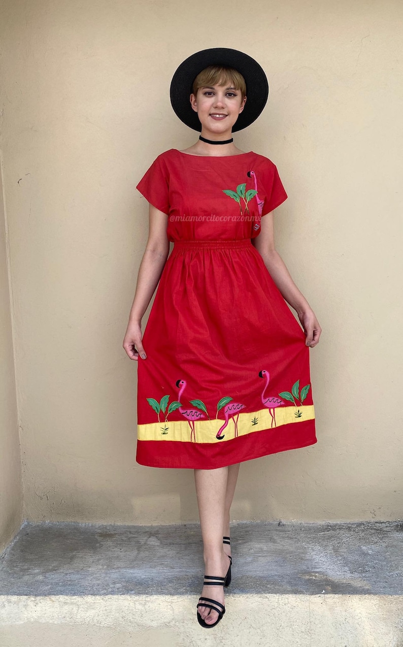 May include: A red midi dress with a gathered waist and short sleeves. The dress features embroidered pink flamingos and green leaves on the skirt. The model is wearing a black hat and black heeled sandals.