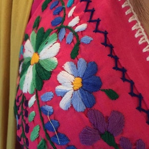 May include: Close-up of a pink fabric with colorful embroidered flowers. The flowers are in shades of blue, green, white, yellow, and purple.