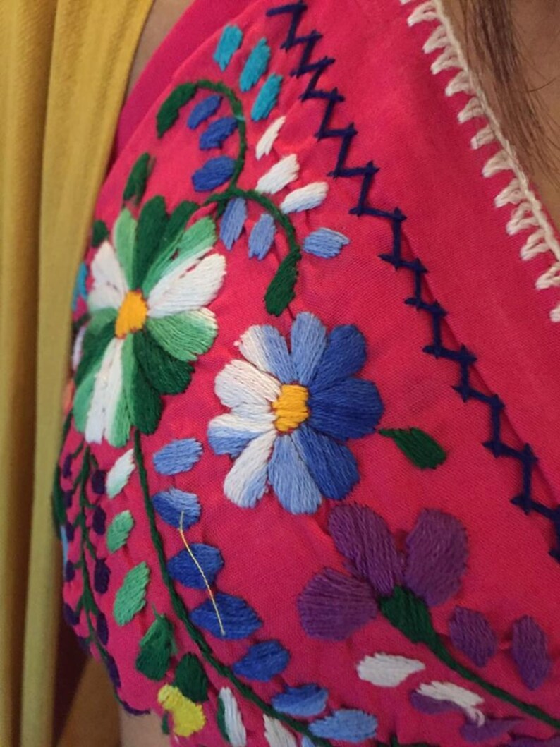 May include: Close-up of a pink fabric with colorful embroidered flowers. The flowers are in shades of blue, green, white, yellow, and purple.