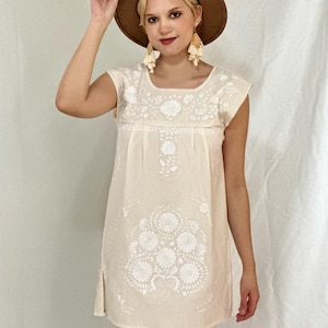 May include: A white cotton dress with white embroidery featuring floral designs. The dress has a square neckline and short sleeves.