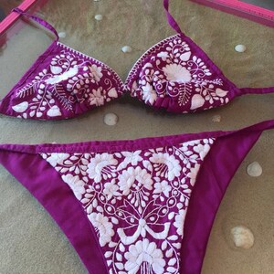 May include: A purple bikini top and bottom with white floral embroidery. The bikini is made of a soft, silky fabric.