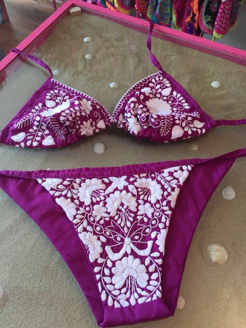 May include: A purple bikini top and bottom with white floral embroidery. The bikini is made of a soft, silky fabric.