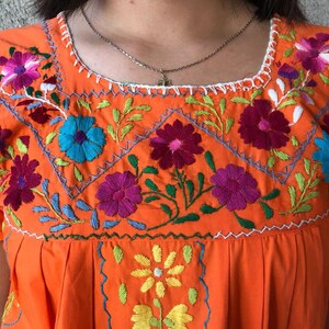 May include: Orange embroidered cotton dress with colorful floral designs. The dress has a round neckline and short sleeves.