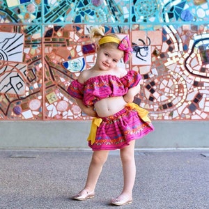 Op de afbeelding: Een jong meisje draagt een roze en gele off-shoulder crop top en een bijpassende rok met bloemenborduursel. Ze staat voor een kleurrijke mozaïekwand.