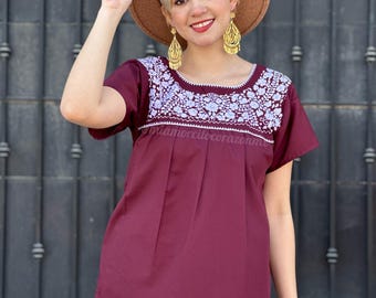 May include: A maroon embroidered blouse with white floral embroidery around the neckline. The blouse has short sleeves and a relaxed fit. The model is wearing a brown hat and gold earrings.