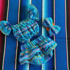 May include: A blue and pink patterned baby outfit with a matching headband. The outfit includes a top with ruffled shoulders and a skirt with ruffles. The headband is a bow with a blue and pink pattern.