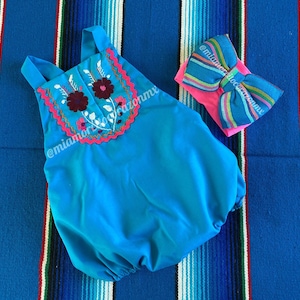 Op de afbeelding: Een turquoise baby onesie met geborduurde bloemen in roze, wit en bordeauxrood. De onesie heeft een gegolfde halslijn en wordt gecombineerd met een roze en blauwe strik.