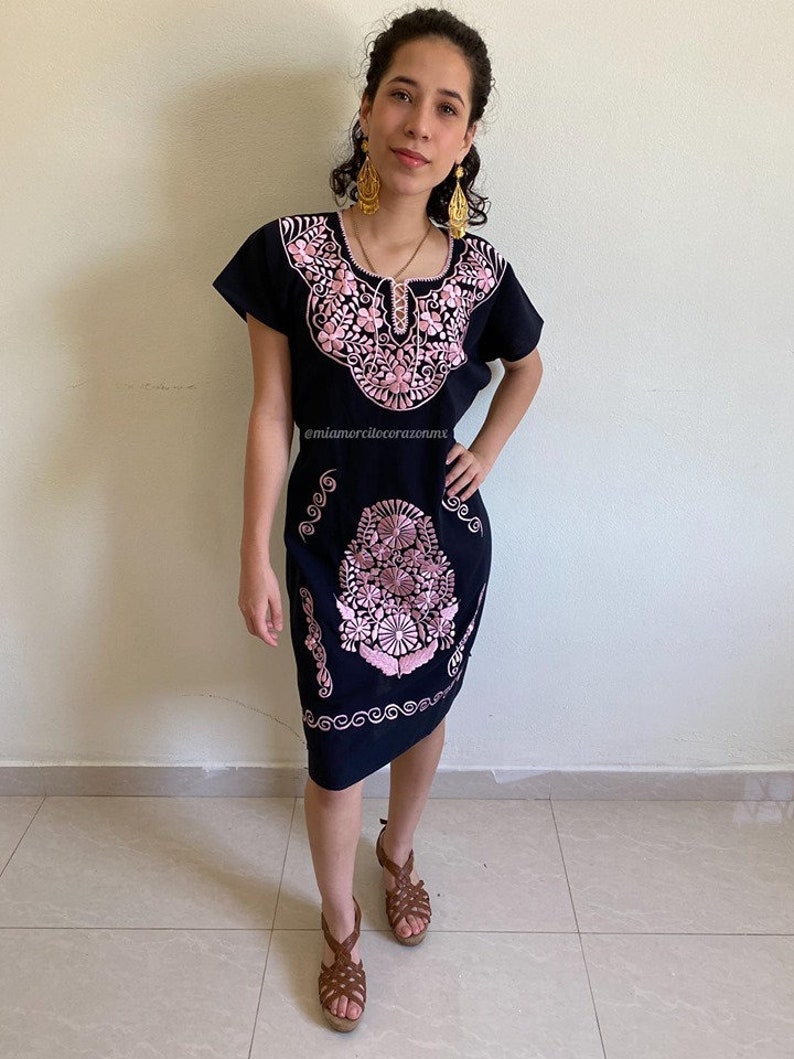 May include: A black embroidered dress with pink floral designs. The dress has short sleeves and a scoop neckline. The model is wearing the dress with brown sandals and gold earrings. The text "@miamorcilcorazonmx" is visible on the dress.