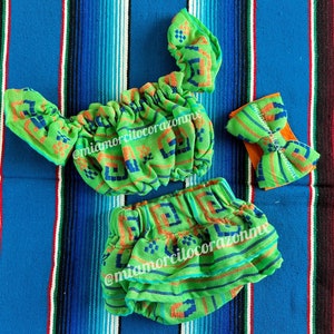 Op de afbeelding: Een groene en oranje outfit voor een baby of peuter, bestaande uit een geruit topje, een broekje en een hoofdband. De outfit heeft een geometrisch patroon en is gemaakt van een zachte, comfortabele stof. De hoofdband is gebonden in een strik. De outfit ligt op een kleurrijke gestreepte achtergrond.