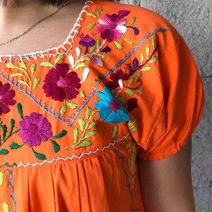 May include: Orange cotton dress with colorful floral embroidery. The embroidery features pink, purple, blue, green, and white flowers.