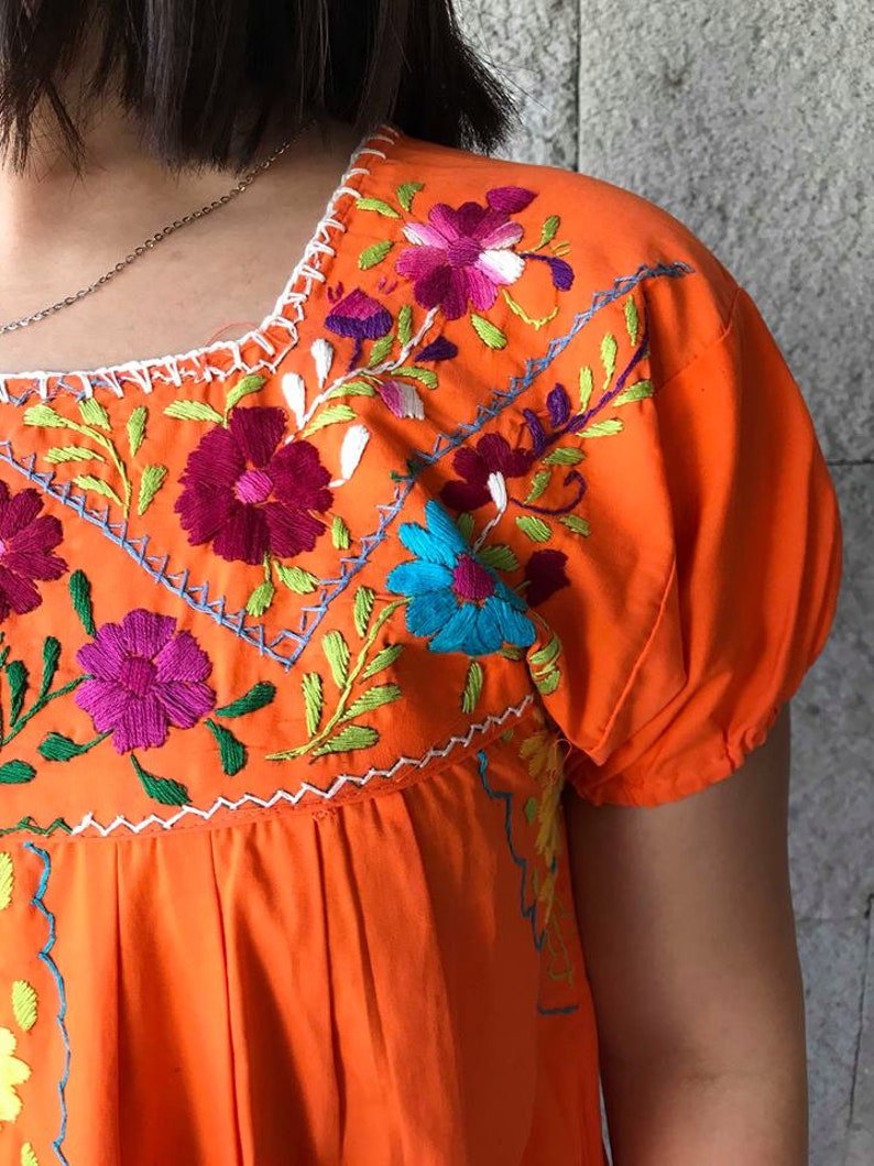 May include: Orange cotton dress with colorful floral embroidery. The embroidery features pink, purple, blue, green, and white flowers.
