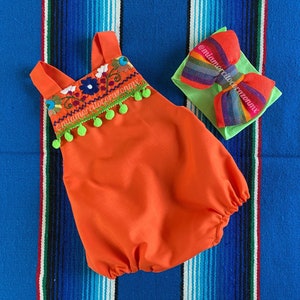 Op de afbeelding: Oranje baby romper met geborduurde bloemen en groene pompoenen. De romper heeft een vierkante halslijn en dunne bandjes. Een groen hoofdband met een veelkleurige strik is inbegrepen. De romper en het hoofdband liggen op een blauw-wit gestreepte achtergrond.