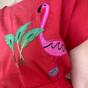 May include: Red cotton dress with a pink flamingo and green leaves embroidered on the front. The dress has a gathered waist and short sleeves.