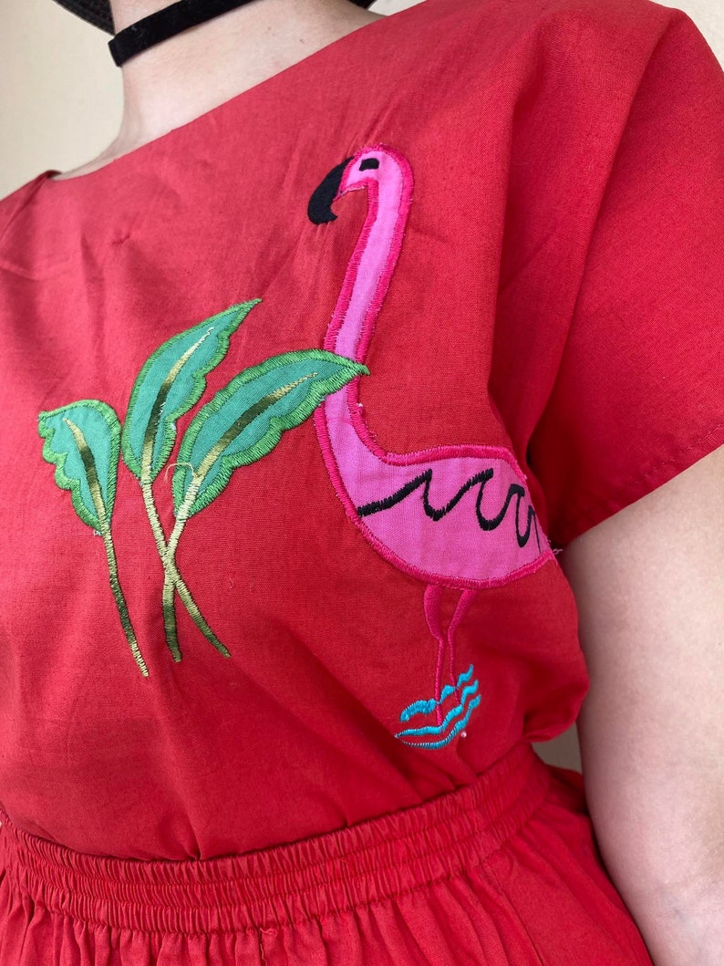 May include: Red cotton dress with a pink flamingo and green leaves embroidered on the front. The dress has a gathered waist and short sleeves.