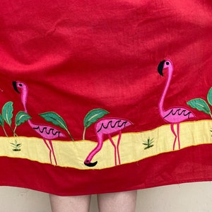 May include: Red cotton skirt with embroidered pink flamingos and green palm trees. The skirt has a yellow band with a white outline.
