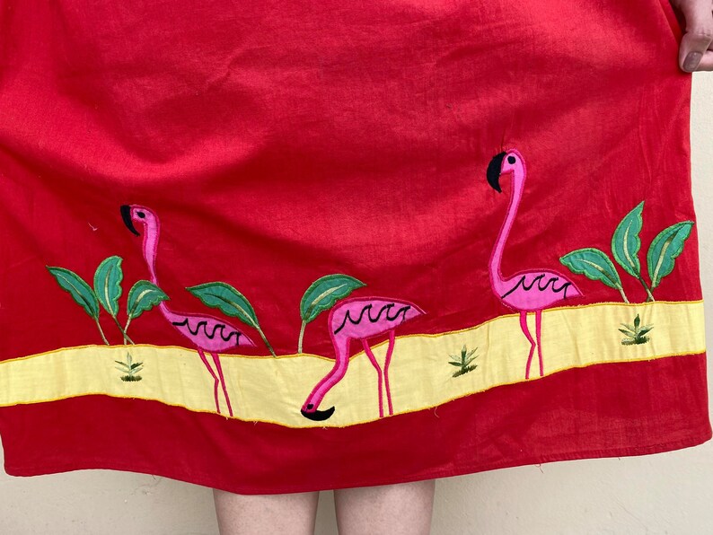 May include: Red cotton skirt with embroidered pink flamingos and green palm trees. The skirt has a yellow band with a white outline.