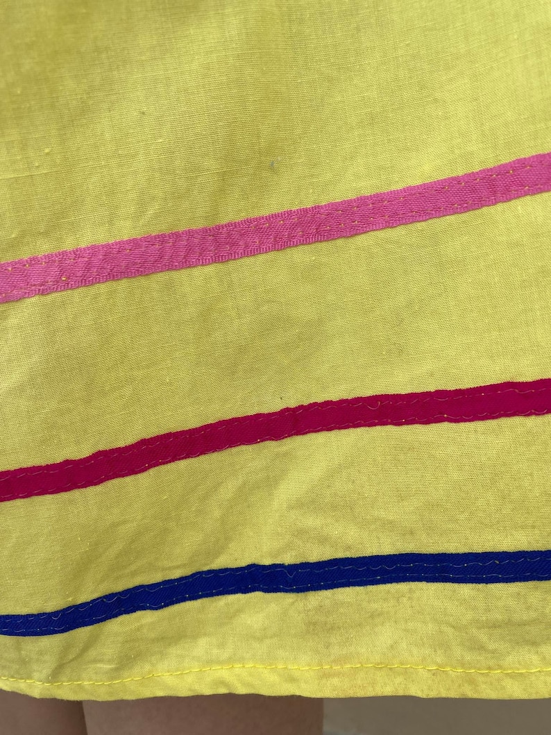 May include: Yellow fabric skirt with pink, red, and blue trim. The skirt has a simple design and is made of a lightweight material.