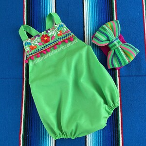 Op de afbeelding: Een groene baby romper met kleurrijke borduursels en pompoentjes rond de halslijn. Een gestreept strikband is ook inbegrepen. De romper en het hoofdband liggen op een kleurrijk gestreept kleed.
