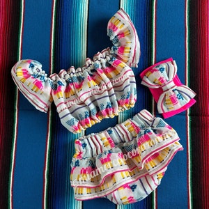 Op de afbeelding: Een witte, roze en gele baby-outfit met een geruite rok en een bijpassend hoofdband. De outfit heeft een geometrisch patroon met lama-ontwerpen. De outfit ligt op een kleurrijk gestreept deken.