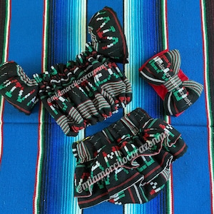 May include: A black and green outfit for a baby or toddler, featuring a ruffled skirt, a top with off-the-shoulder sleeves, and a headband with a bow. The fabric has a Mexican-inspired pattern with red, white, and green stripes. The outfit is laid out on a colorful striped blanket.