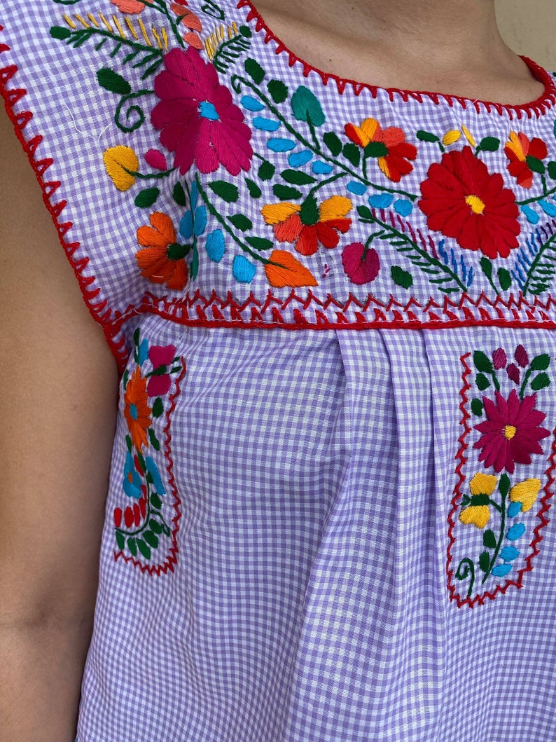 Peut inclure: Une blouse sans manches en vichy violet et blanc avec des fleurs brod&eacute;es color&eacute;es autour de l'encolure.