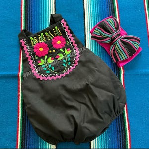 Op de afbeelding: Zwarte babyromper met bloemenborduurwerk in roze, groen en geel. De romper heeft een roze geschulpte rand en een bijpassende strik-hoofdband met gestreepte patronen in zwart, roze, groen en blauw. De items worden gepresenteerd op een kleurrijk gestreept textiel.