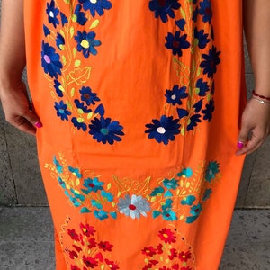 May include: Orange cotton maxi dress with colorful floral embroidery. The dress features a round neckline and a loose, flowing silhouette.