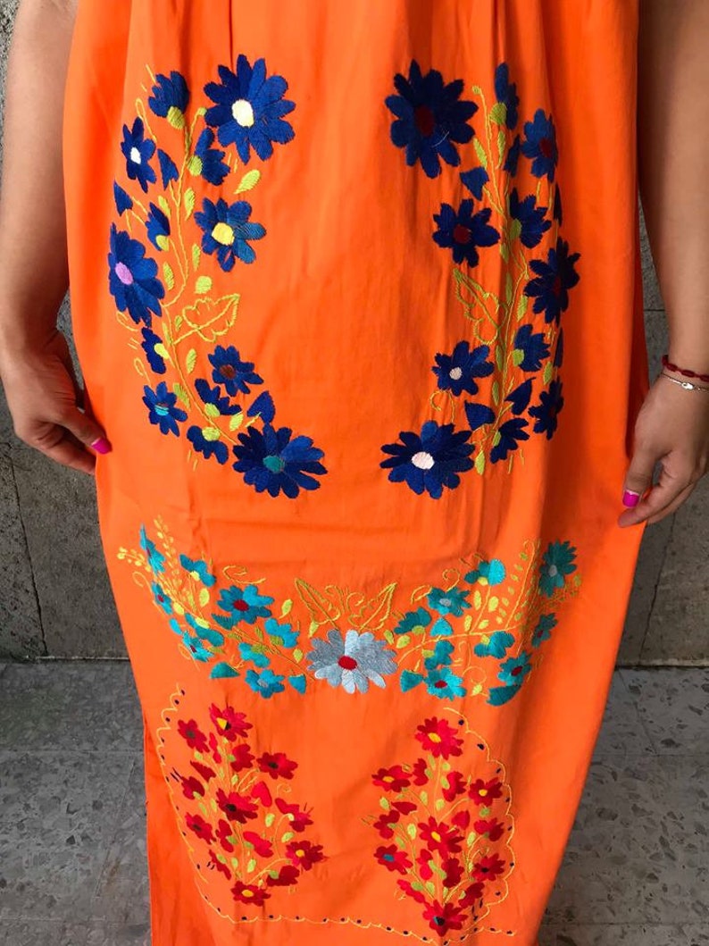 May include: Orange cotton maxi dress with colorful floral embroidery. The dress features a round neckline and a loose, flowing silhouette.