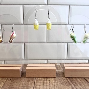 May include: Three pairs of dangle earrings displayed on clear acrylic stands with wooden bases. The earrings feature miniature food items, including milkshakes, ice cream, and green drinks.