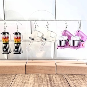 May include: Three pairs of kitchen appliance earrings. The first pair is silver blenders with colorful fruit inside. The second pair is white stand mixers with silver bowls. The third pair is pink stand mixers with silver bowls. The earrings are displayed on a wooden stand.