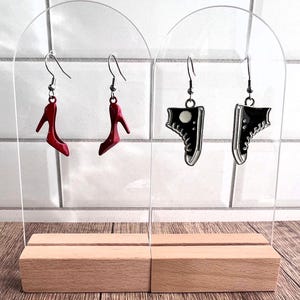 May include: Two pairs of earrings on a clear acrylic display stand. One pair features red high heel shoes and the other pair features black sneakers.
