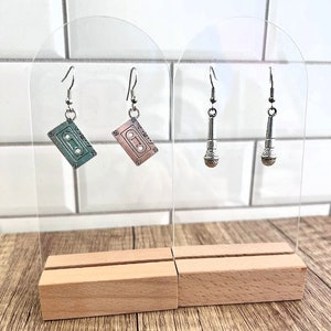 May include: Two pairs of earrings on a wooden display stand. One pair features a silver cassette tape charm, the other pair features a silver microphone charm.