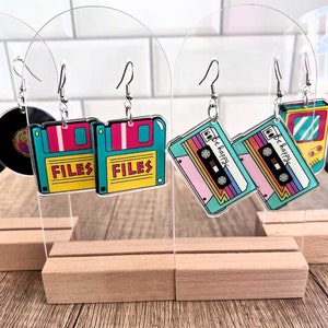May include: Assortment of colorful retro-themed earrings featuring designs like a floppy disk, cassette tape, and a video game console. The earrings are displayed on clear acrylic stands.