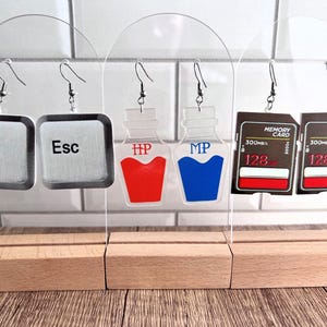 May include: Six pairs of earrings on display stands. The earrings are made of clear acrylic and feature different designs, including a computer key labeled "Esc", a potion bottle with "HP" written on it, a potion bottle with "MP" written on it, and a memory card with "MEMORY CARD 300MB 128" written on it.