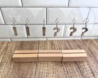 May include: Three acrylic earring display stands with silver earrings. The stands are on a wooden surface. The earrings are in the shape of a question mark and a rectangle.