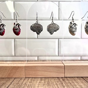 May include: Three pairs of dangle earrings displayed on a wooden stand. The earrings feature silver heart and brain charms. The hearts are red with silver detailing. The brains are silver.