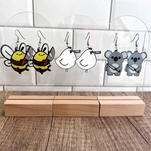 May include: Three sets of earrings displayed on acrylic stands. The first set features two yellow and black bee earrings holding a small axe. The second set features two white chicken earrings holding a knife. The third set features two gray koala earrings holding a knife.