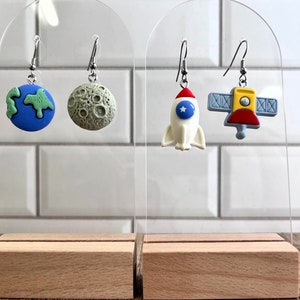May include: A set of four space-themed earrings. The earrings are made of polymer clay and feature a blue and green Earth, a gray moon, a white rocket with a red nose and a blue star, and a yellow satellite with a red nose.