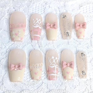 Può includere: Un set di dieci unghie finte con vari disegni. Le unghie sono principalmente beige chiaro, con dettagli rosa e bianchi. Alcune unghie presentano fiocchi, cuori e glitter. Altre hanno motivi intricati.
